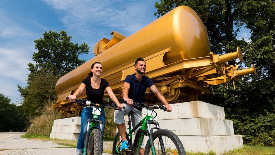 Zwei Personen fahren mit Fahrrädern vor einer goldenen Skulptur im Österreichischen Skulpturenpark. | © Region Graz - Harry Schiffer